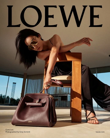Greta Lee wearing sunglasses and black clothing, leaning over a wooden chair while holding a burgundy leather handbag in a bright interior.
