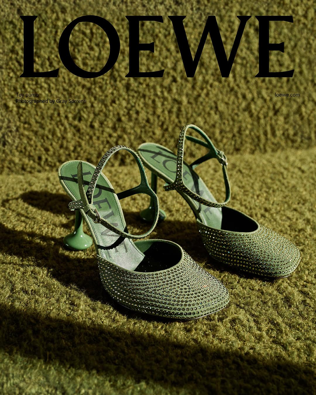 Pair of jeweled green slingback heels resting on a textured olive green surface for Loewe campaign.