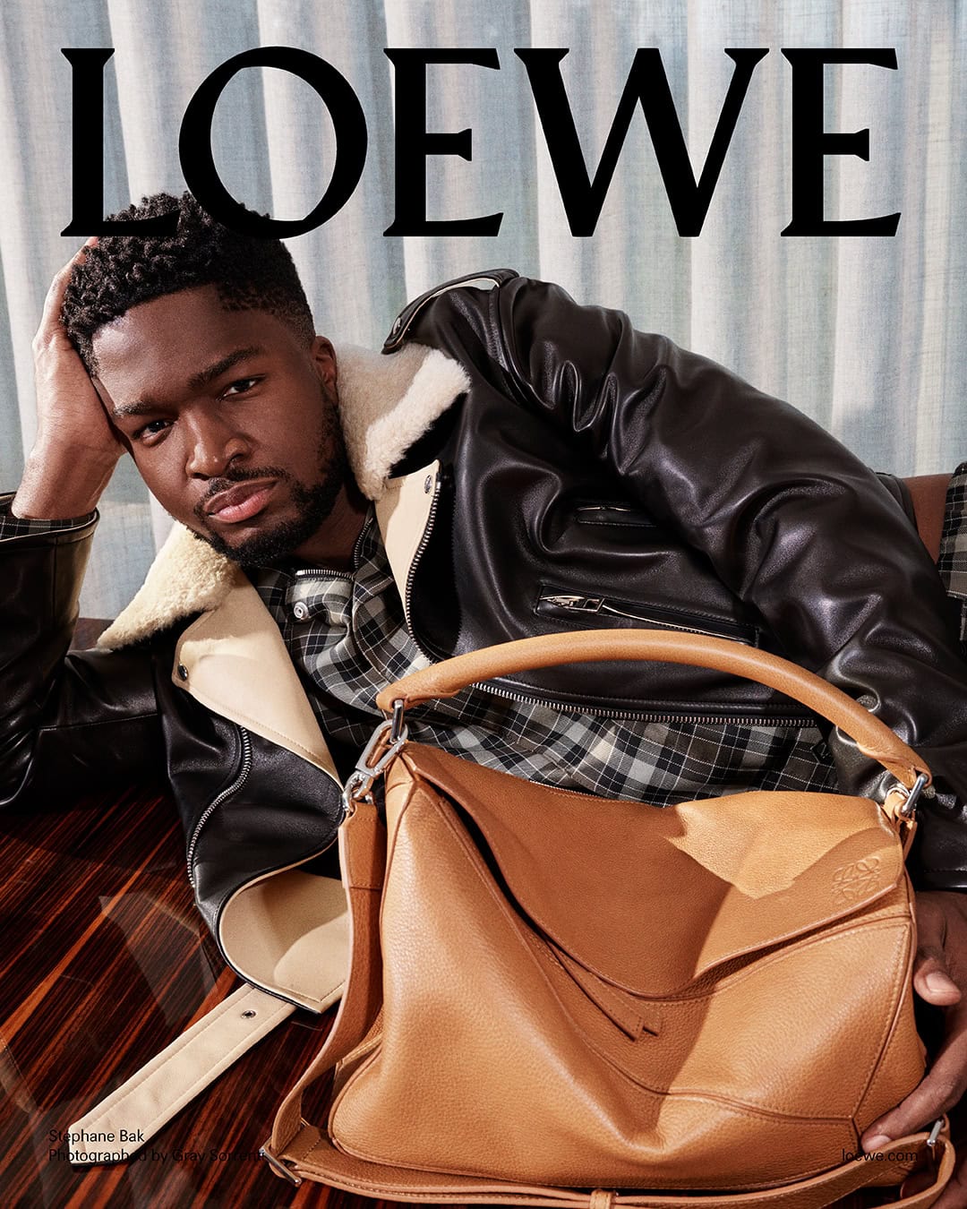 Portrait of actor Stéphane Bak wearing a black shearling jacket and plaid shirt, holding a tan leather bag.