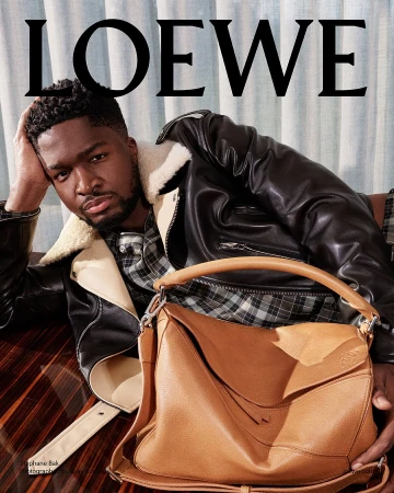 Portrait of actor Stéphane Bak wearing a black shearling jacket and plaid shirt, holding a tan leather bag.