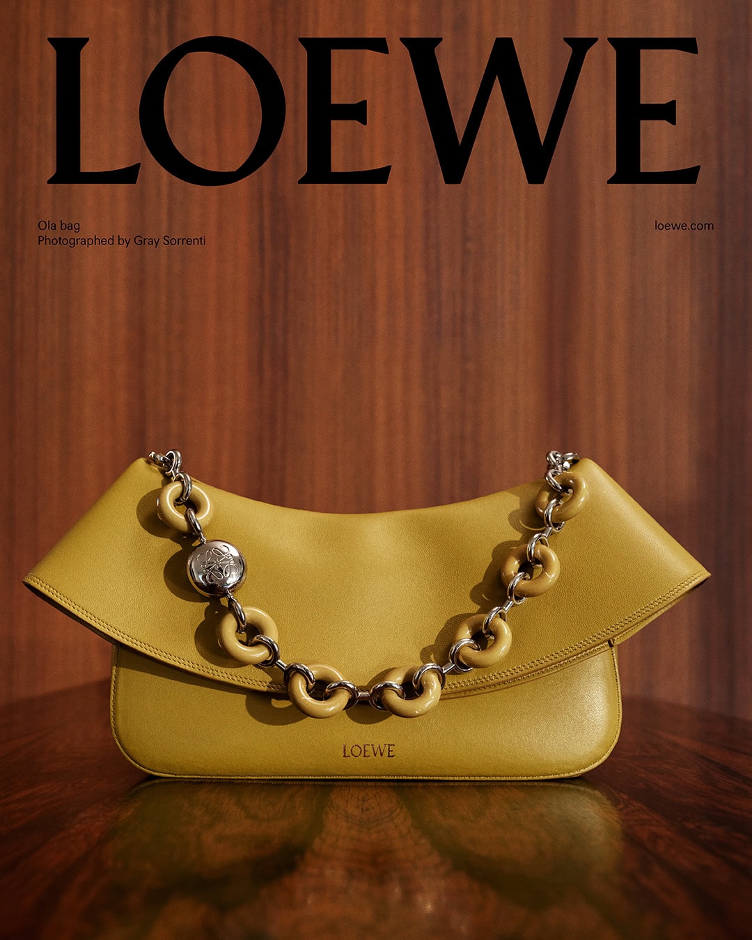 Yellow leather shoulder bag with chain strap centered against a rich wood panel background under Loewe branding