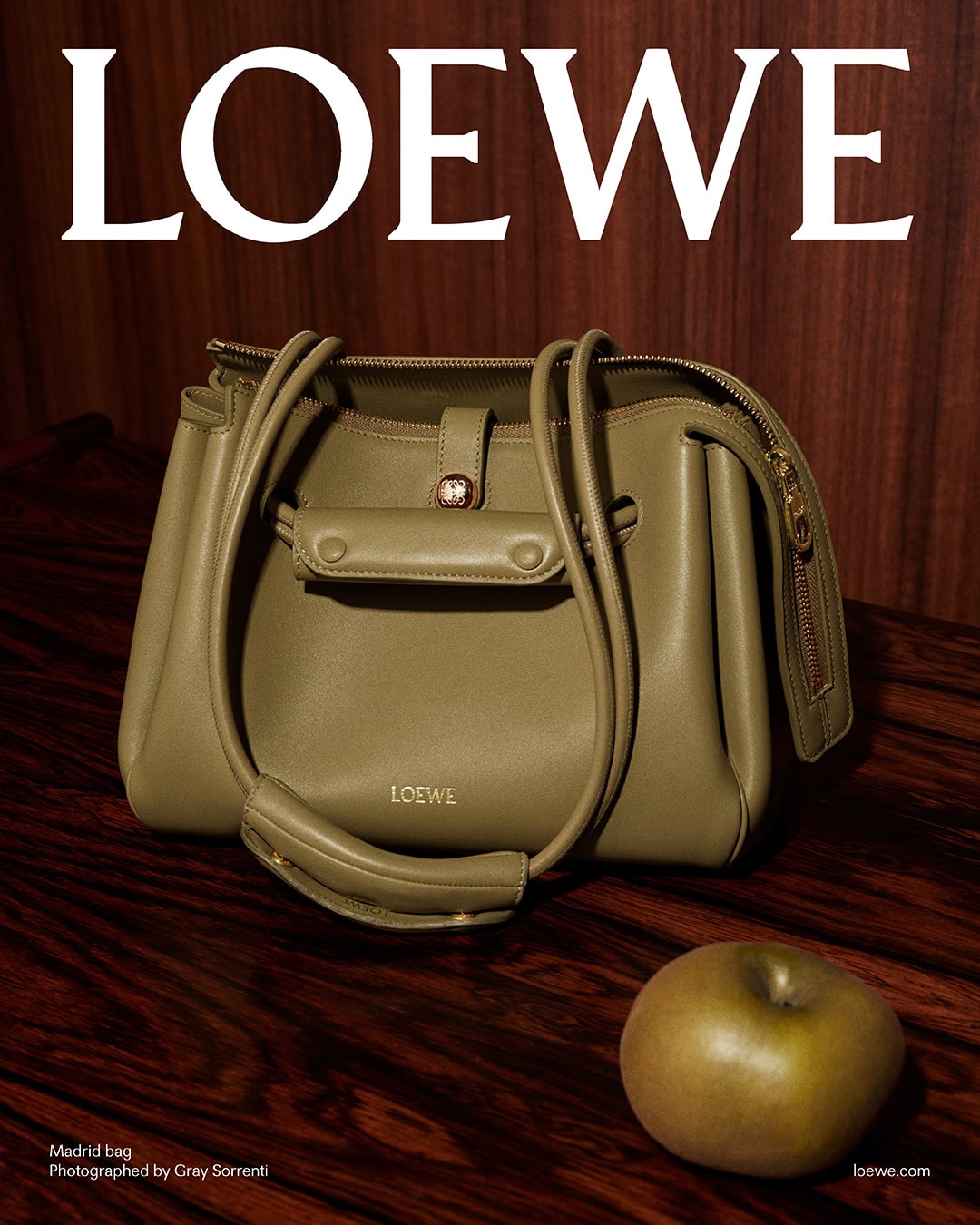 Olive green leather Loewe handbag resting on a polished dark wood table next to a brownish apple