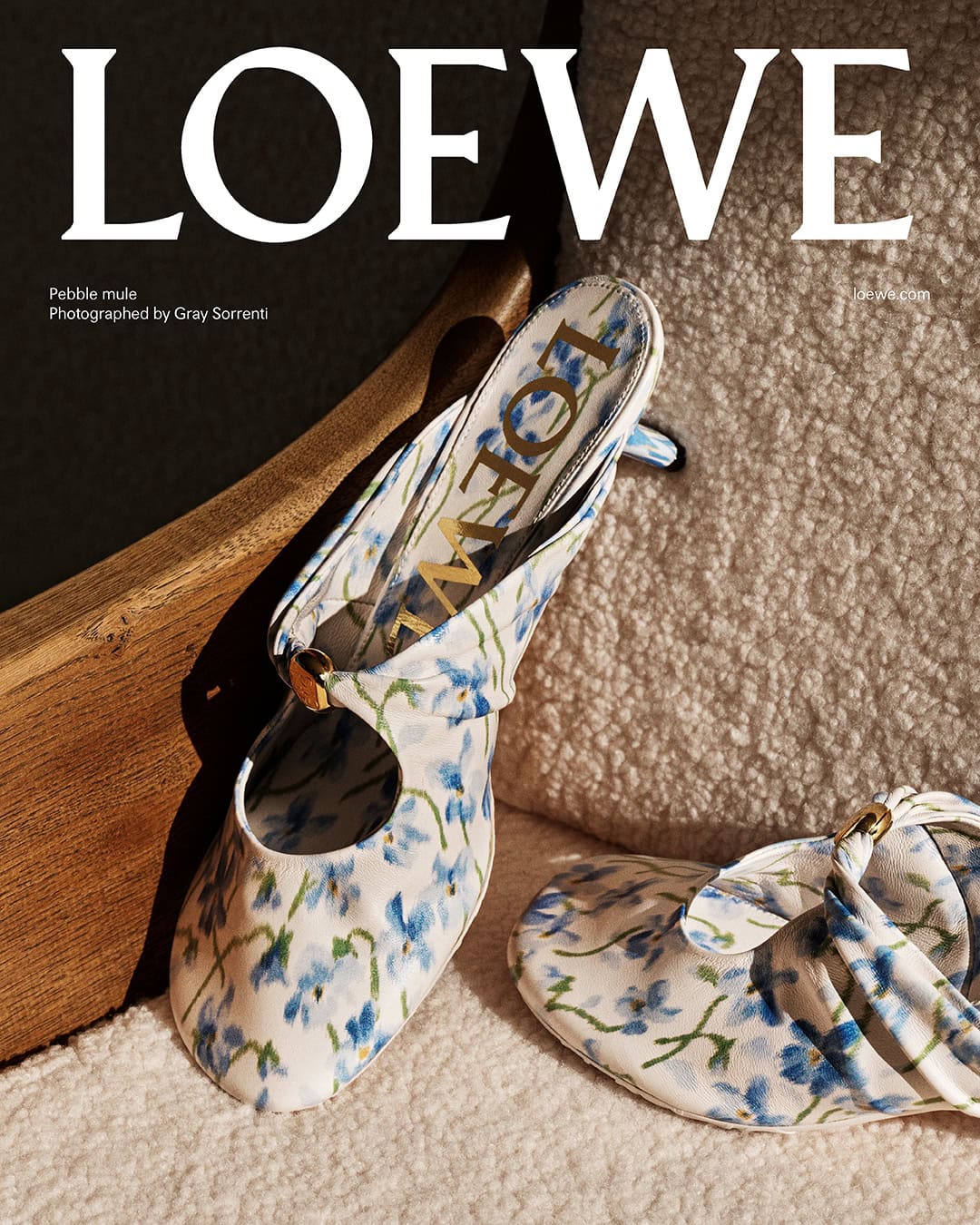 Loewe floral print mules resting on a textured white fluffy cushion next to a wooden frame