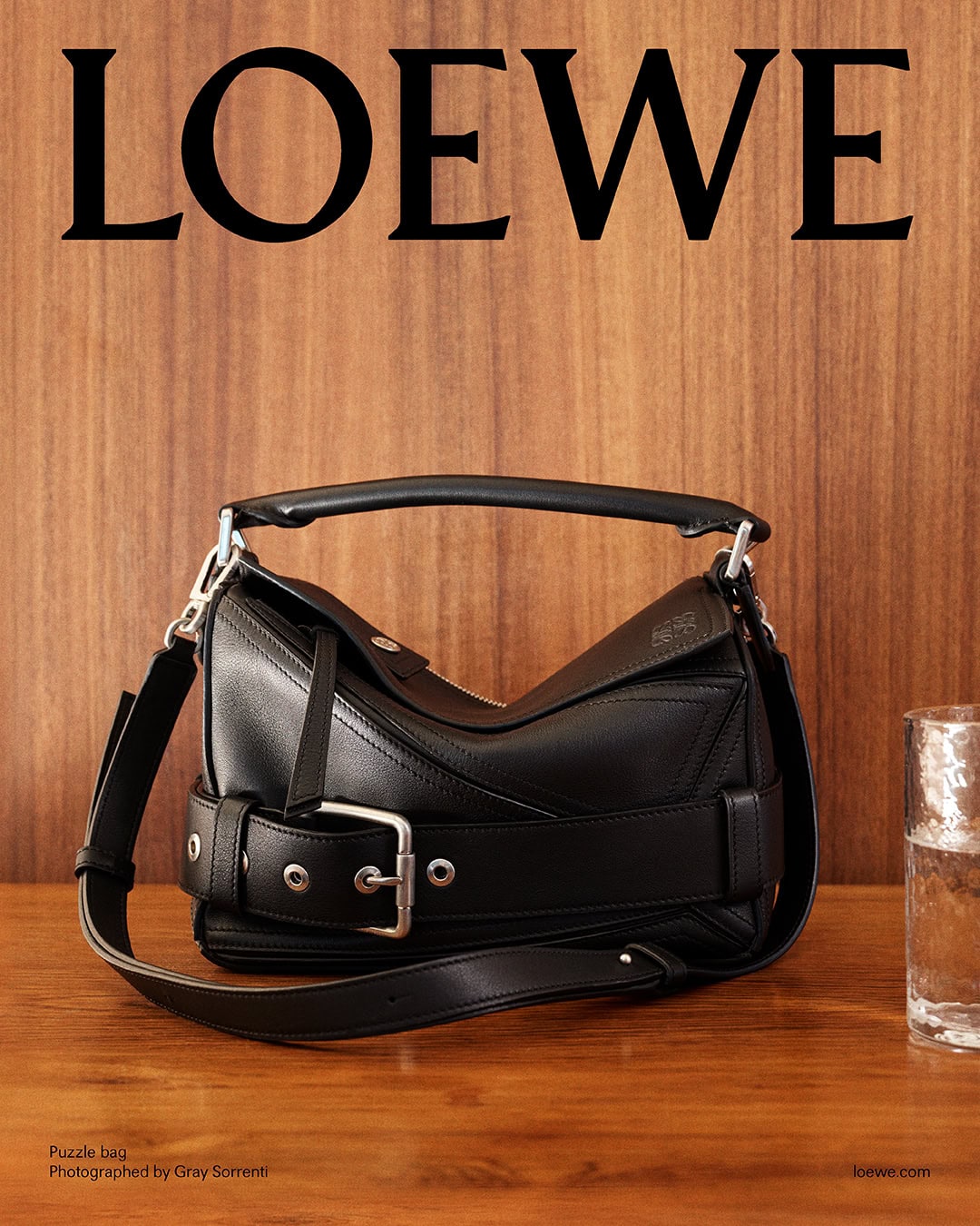 Black leather Puzzle bag resting on a wooden surface against a vertical wood panel wall with large LOEWE branding.