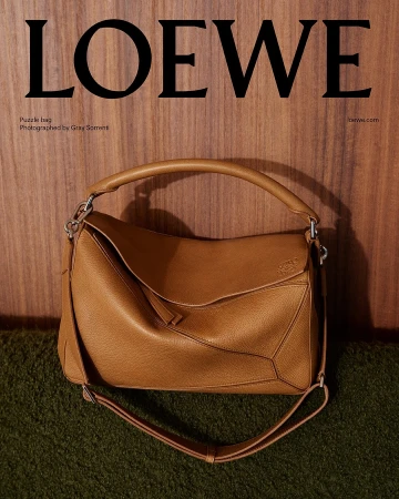Tan leather Loewe puzzle bag resting against a vertical wood panel wall above a textured green surface.