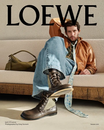 Josh O Connor seated on a textured beige sofa wearing a brown leather jacket and boots for Loewe Pre-Fall 2025 campaign