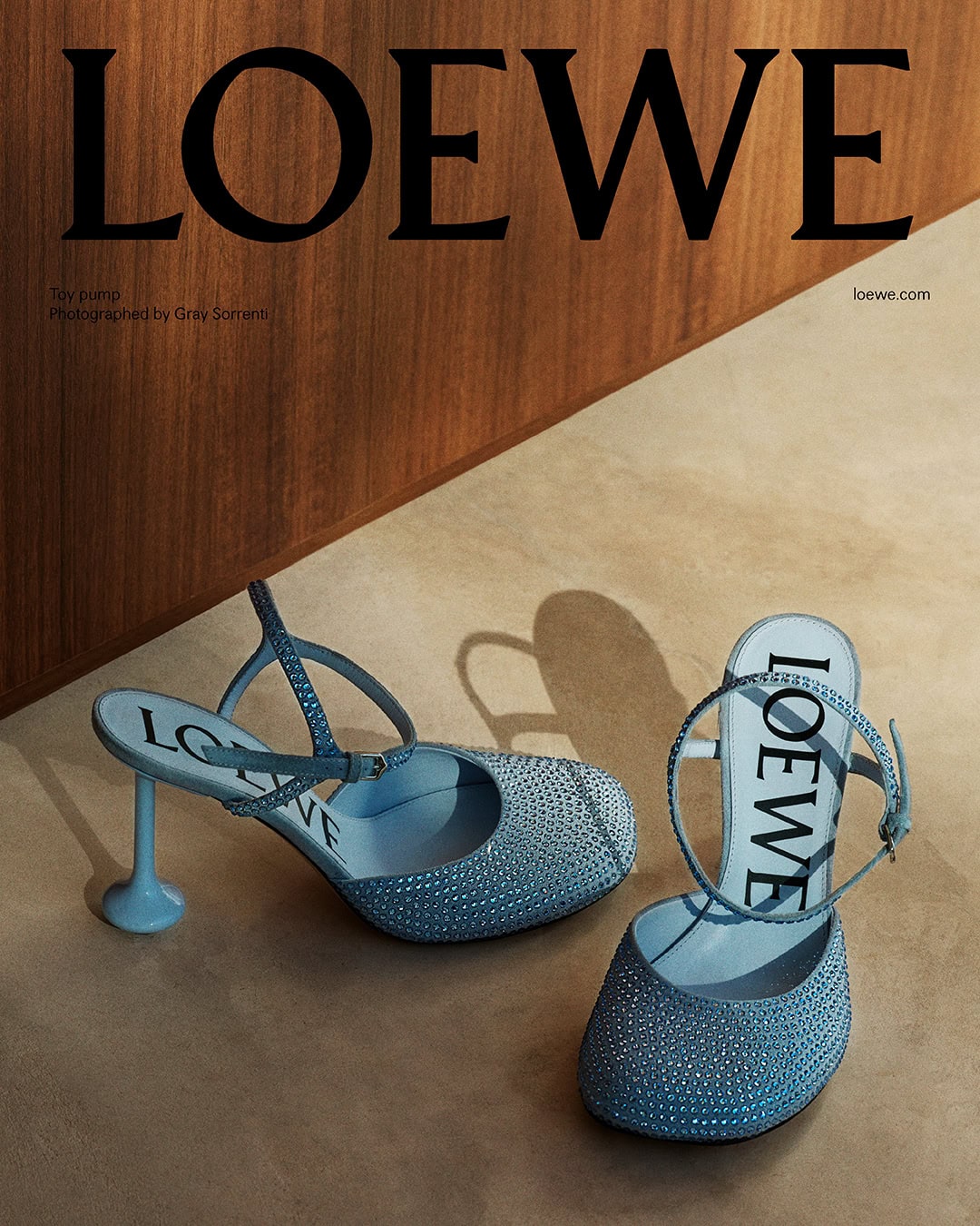 Pair of light blue embellished pumps with slingback straps placed on a warm beige surface against a wooden wall.