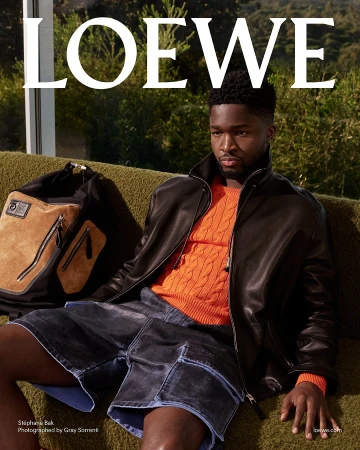 Stéphane Bak seated on a green textured sofa wearing an orange knit sweater and black leather jacket next to a large duffel bag