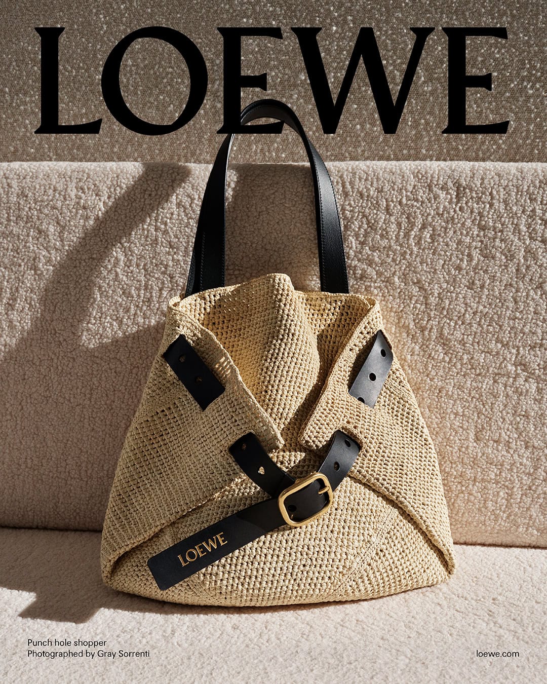 Loewe straw shopper bag with black leather straps and gold buckle against a textured fabric background