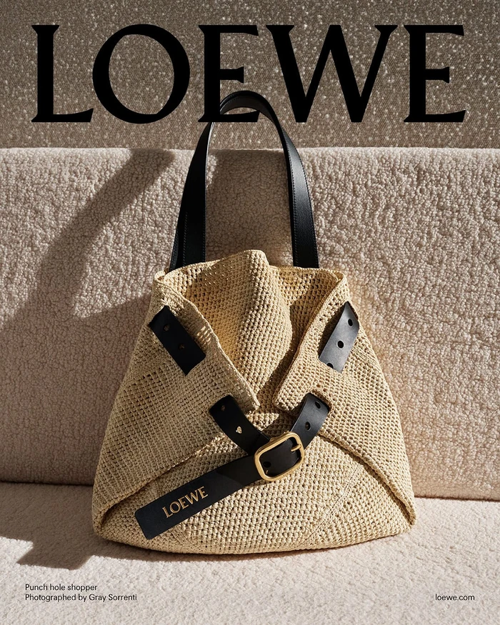 Loewe straw shopper bag with black leather straps and gold buckle against a textured fabric background