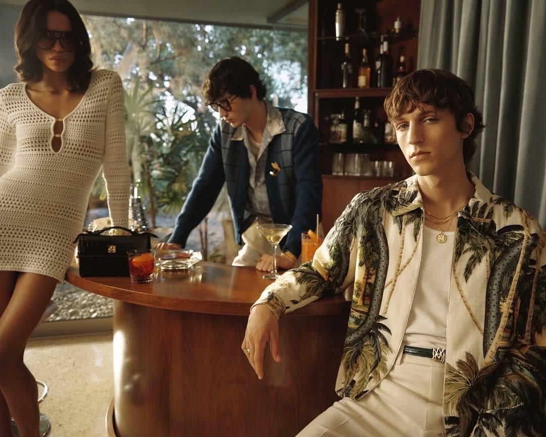 Three models in vintage attire gathered around a wooden bar inside a mid-century home interior.