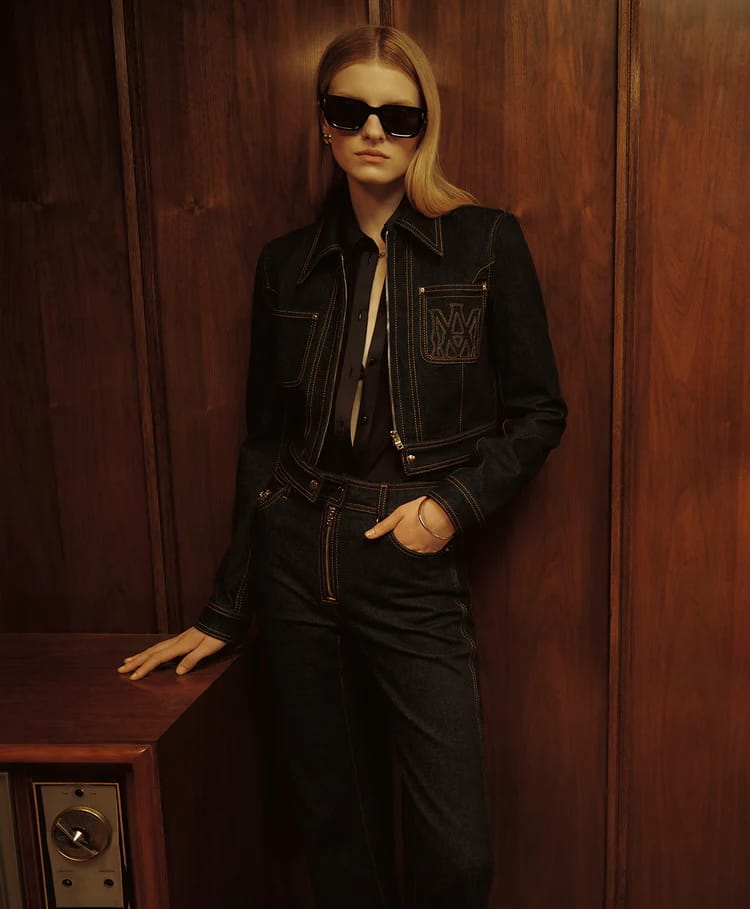 Model in dark sunglasses and a dark denim jacket standing against wood paneling for Amiri Pre-Fall 2025