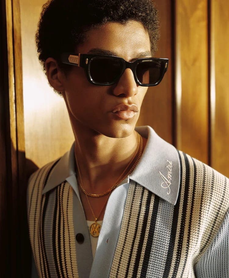Close-up portrait of a Black man wearing black sunglasses, gold necklaces, and a light blue and white striped cardigan against wood paneling
