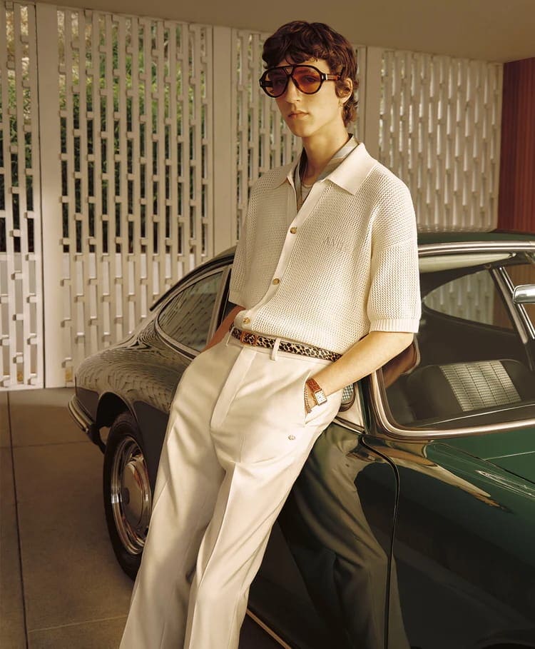 A man in a cream knit polo shirt and sunglasses leans against a vintage green car in a carport.
