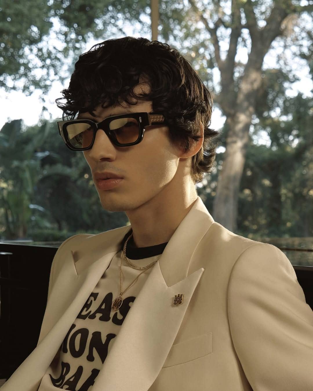 Man wearing sunglasses and light blazer outdoors against a background of trees