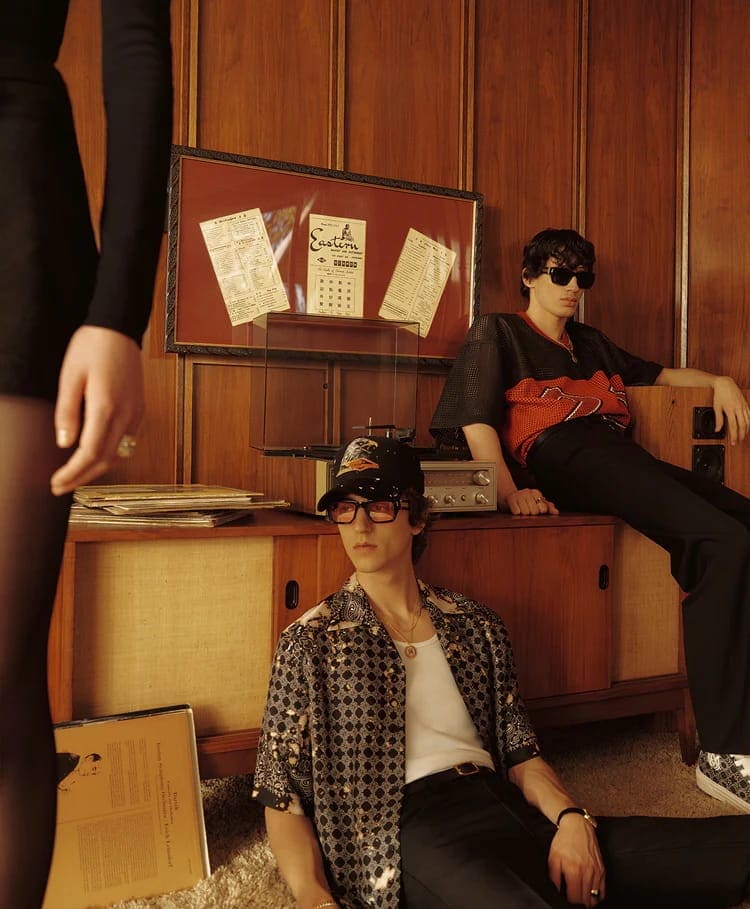 Two young male models in retro attire relaxing inside a wood-paneled room for Amiri Pre-Fall 2025 campaign.