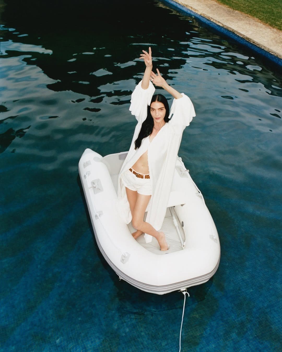 Model standing in a small white inflatable boat floating in a blue swimming pool during a campaign shoot