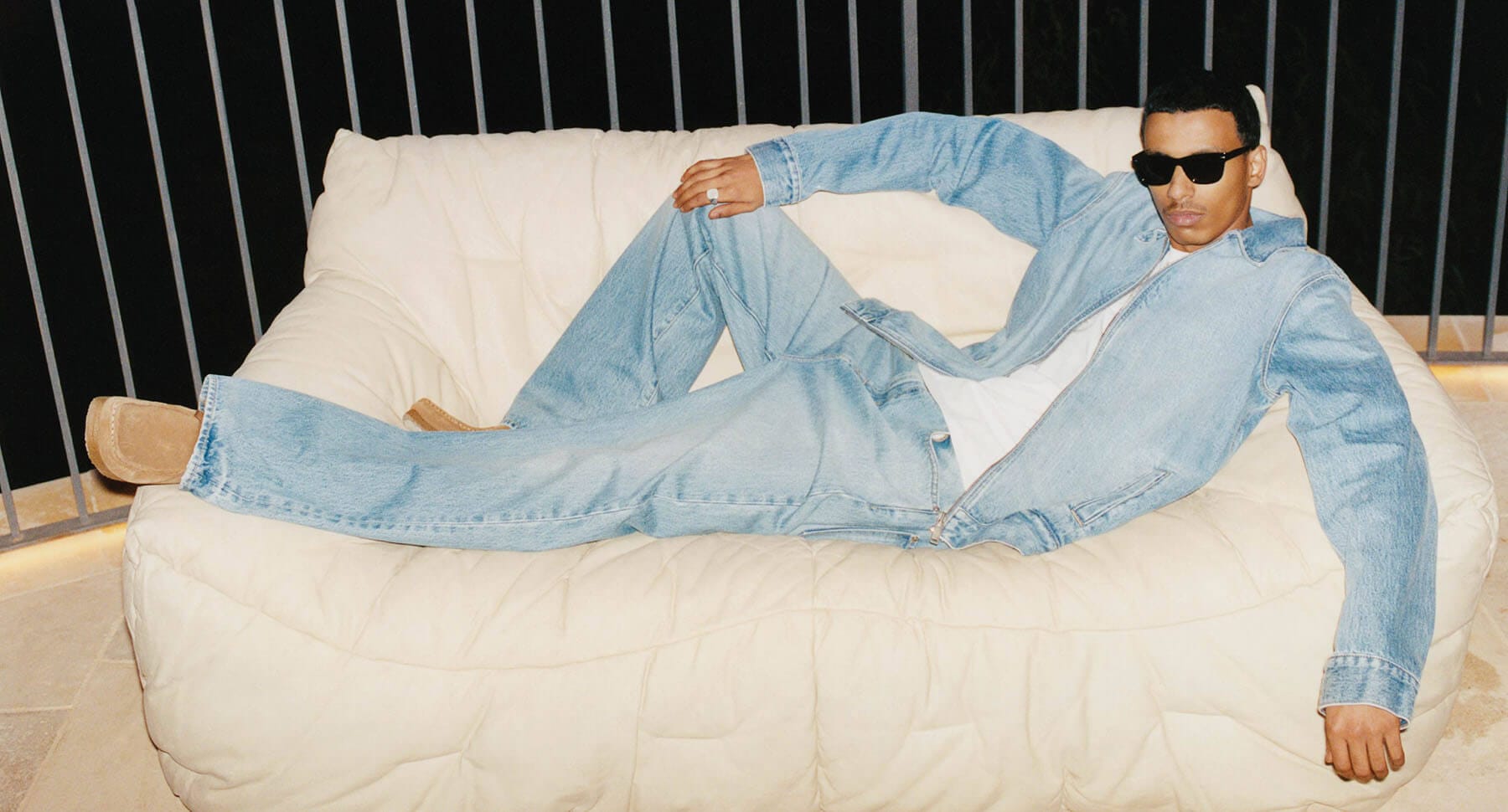 A man wearing a denim jacket and jeans reclining on a cream-colored puffy sofa at night
