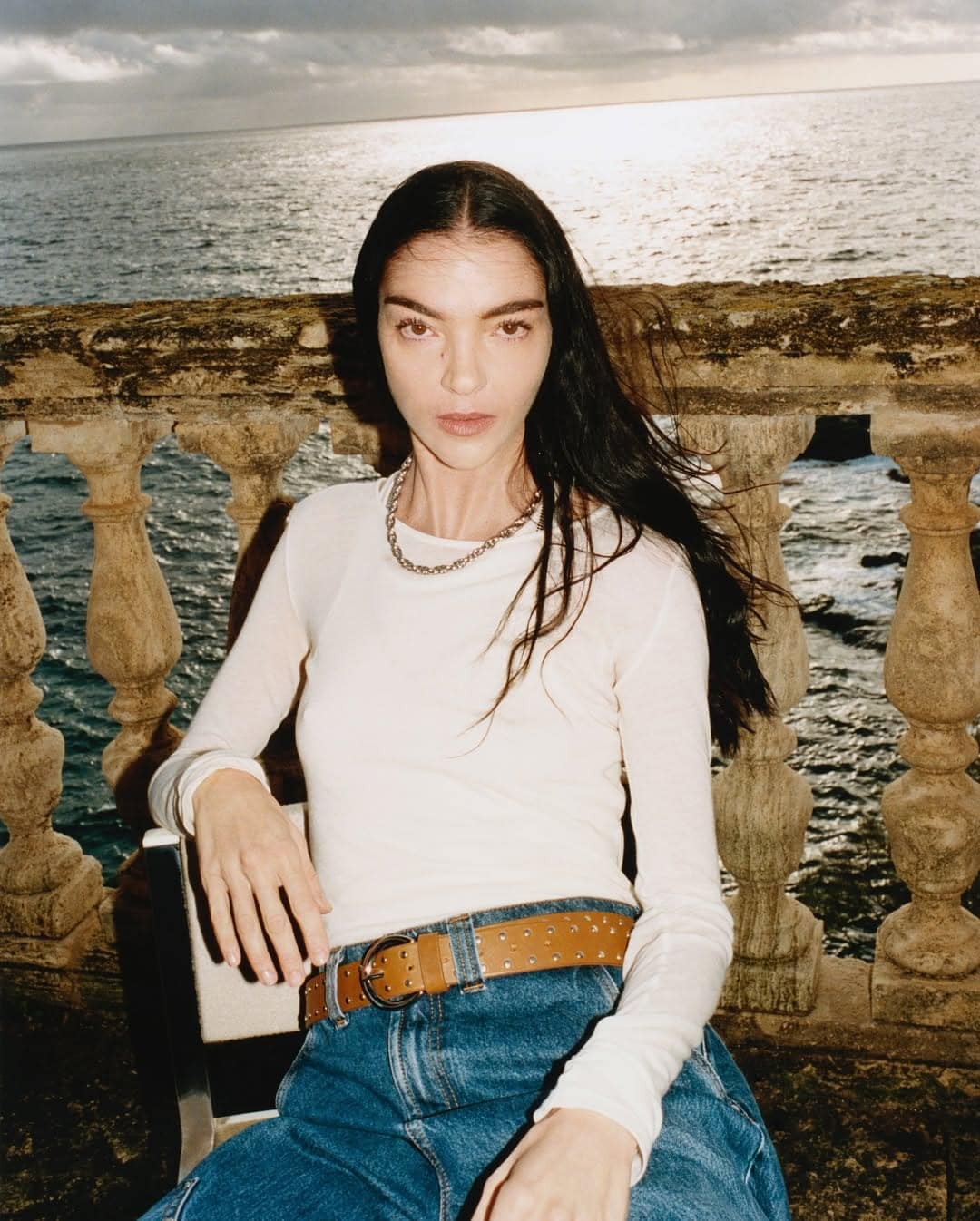 Model seated outdoors by the sea wearing a white long-sleeve top, blue jeans, and a brown studded belt against a stone balustrade.