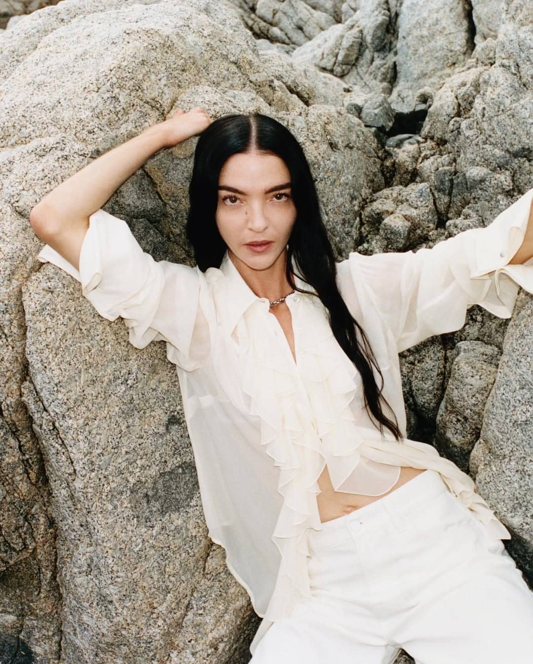 Mariacarla Boscono leaning against coastal rock formation in white blouse and trousers for 7 For All Mankind