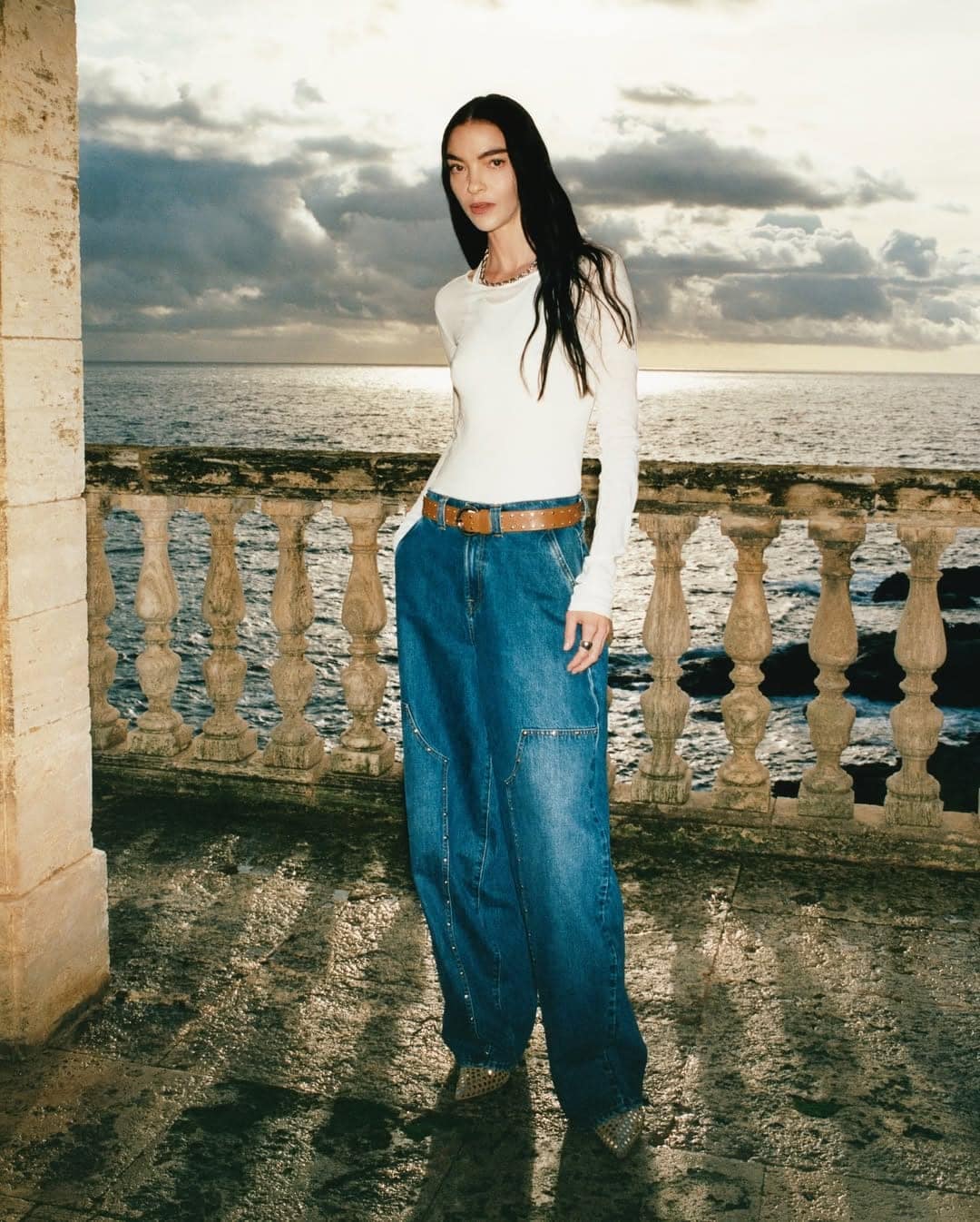 Model standing on a stone balcony by the ocean wearing a white long-sleeve top and wide-leg denim jeans