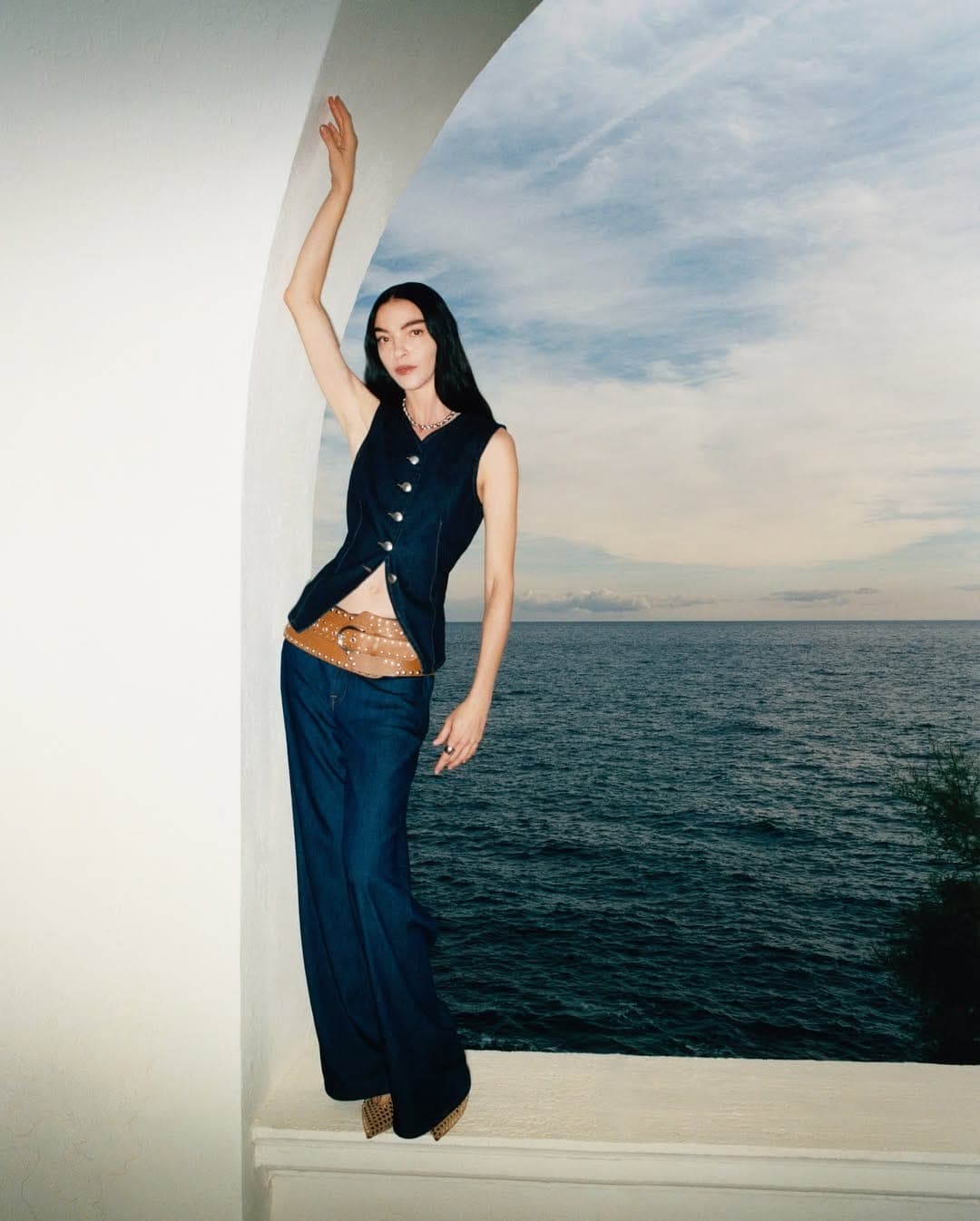 Model standing inside an arched opening by the sea wearing dark denim vest and trousers with a wide brown belt.