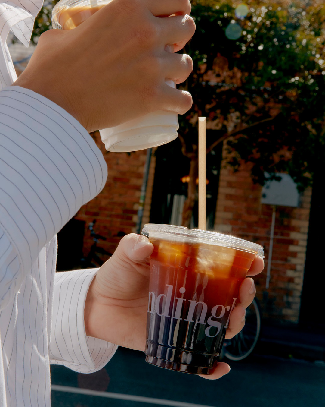 Close-up of hands holding plastic iced coffee cups in front of a brick exterior for Standing Room brand identity
