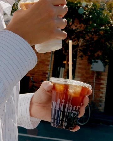 Close-up of hands holding plastic iced coffee cups in front of a brick exterior for Standing Room brand identity