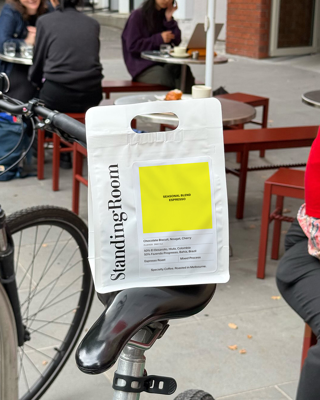 White Standing Room coffee bag with a yellow label perched on a bicycle seat on an outdoor street