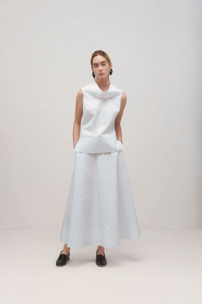 Elegant woman in a sleeveless white top and long skirt posing in a studio, complemented by dark flats and minimal jewelry. 