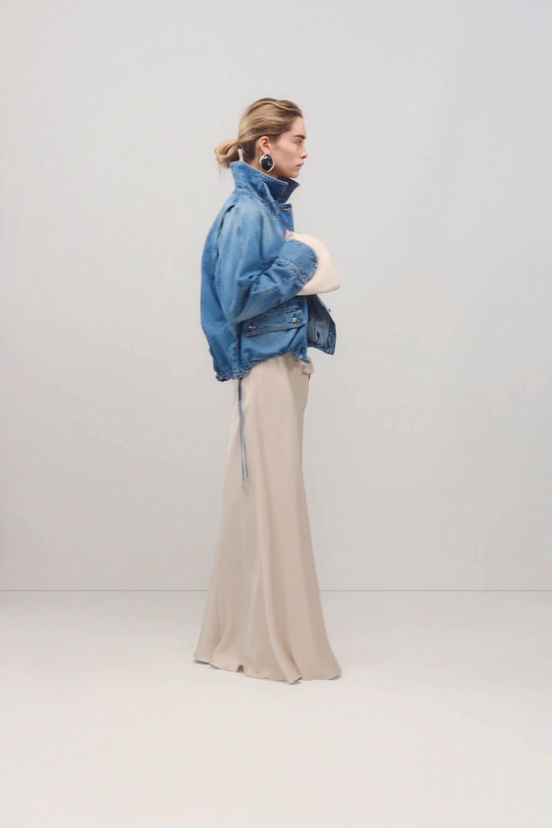 Fashionable woman in denim jacket and long skirt strikes a pose in a bright, minimalist studio setting, showcasing contemporary style.