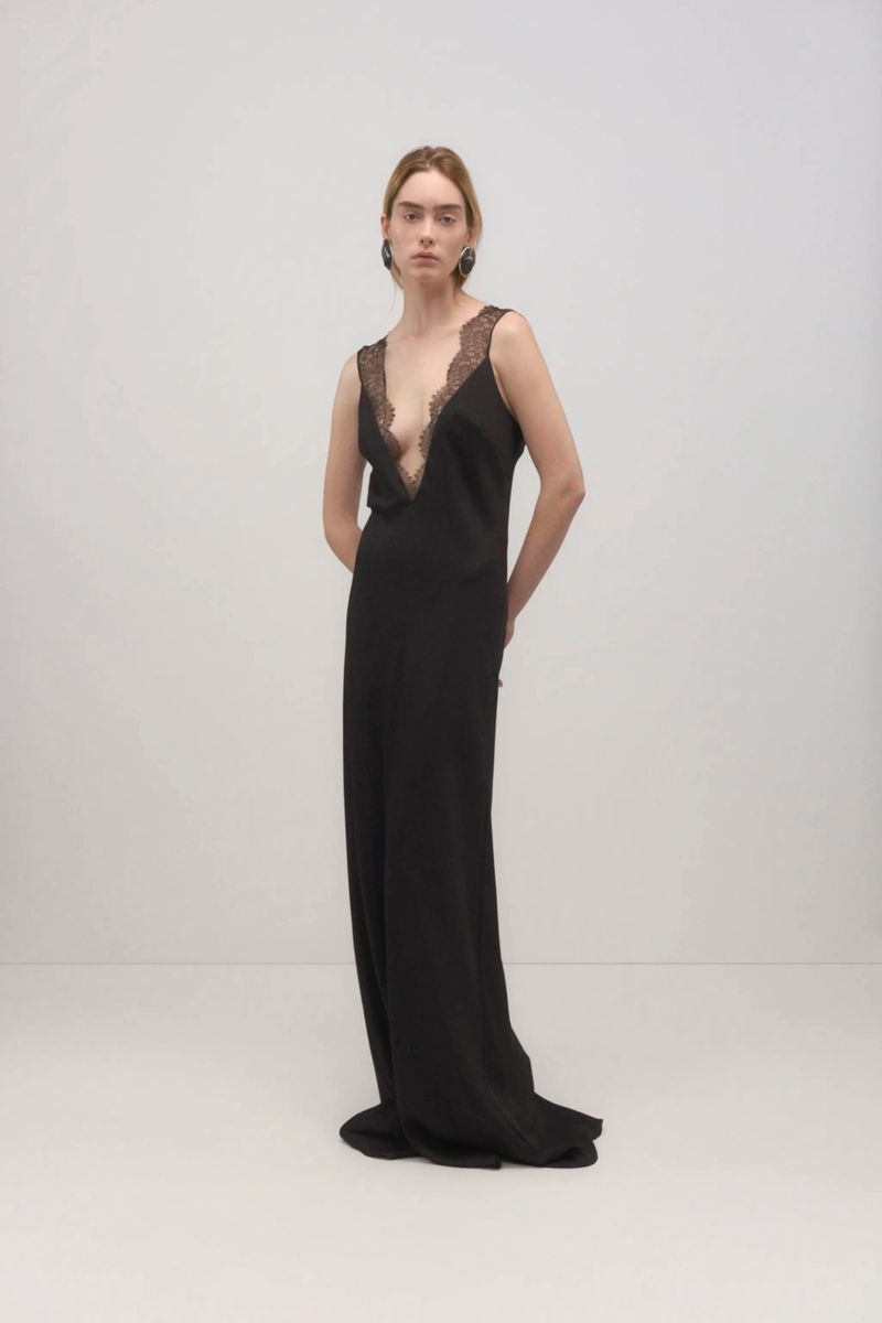 A poised adult woman in a black sleeveless gown with lace trim, indoor studio lighting, neutral background, fashionable earrings.
