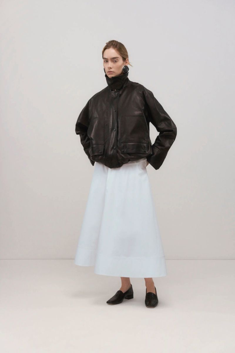 Studio shot of a woman in a dark leather jacket and white skirt. The model poses against an off-white background with neutral lighting.