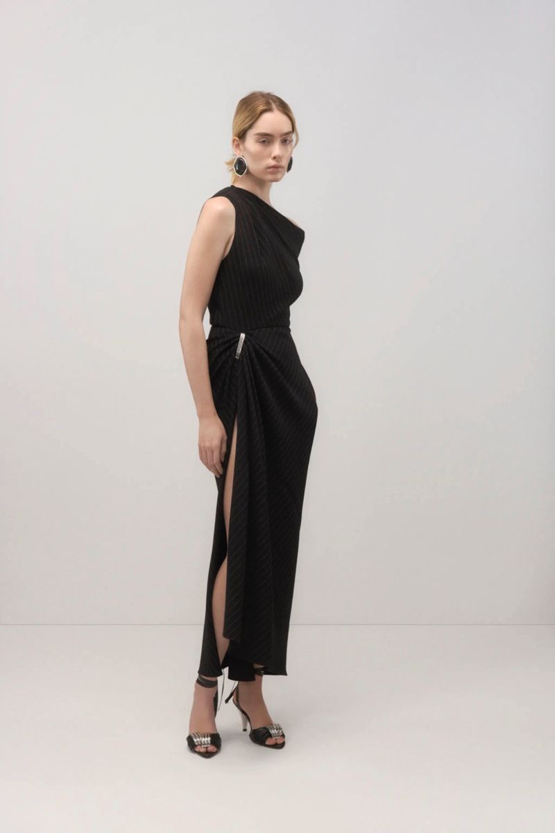 A woman in a black sleeveless dress with a high slit and black high heels poses in a studio against a light background.