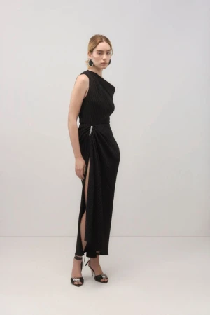 A woman in a black sleeveless dress with a high slit and black high heels poses in a studio against a light background.