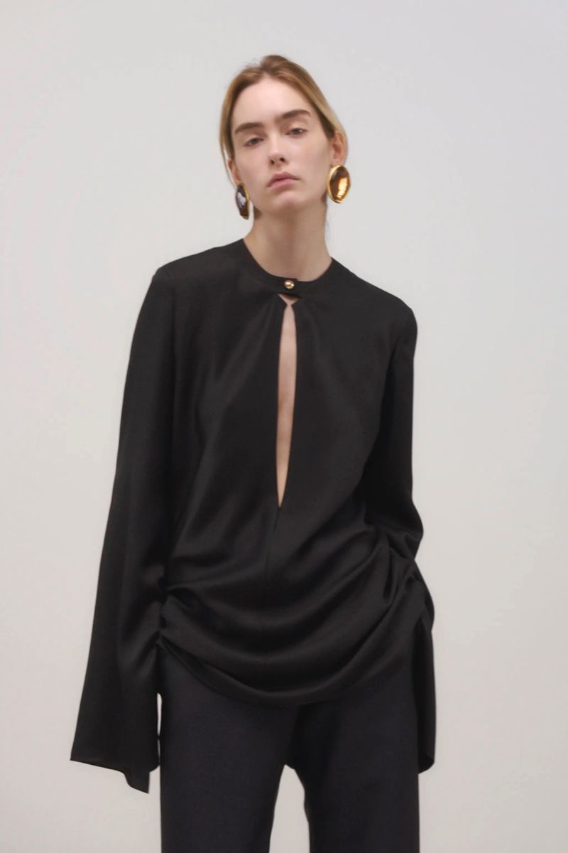 A poised woman in a black blouse and pants stands against a neutral backdrop, accessorized with prominent gold earrings for a fashion-forward look.
