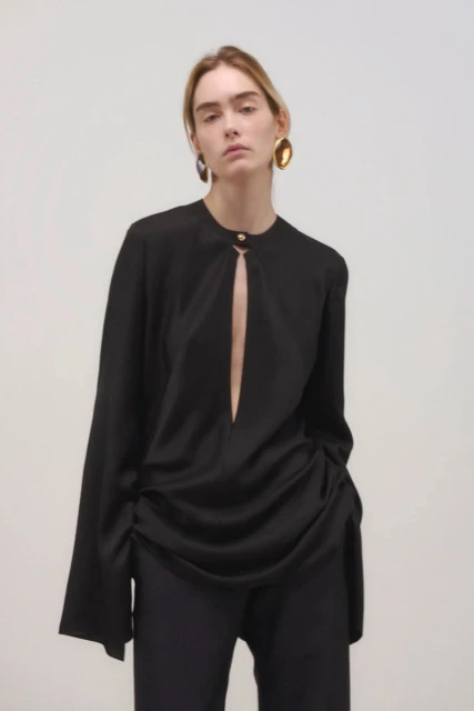 A poised woman in a black blouse and pants stands against a neutral backdrop, accessorized with prominent gold earrings for a fashion-forward look.