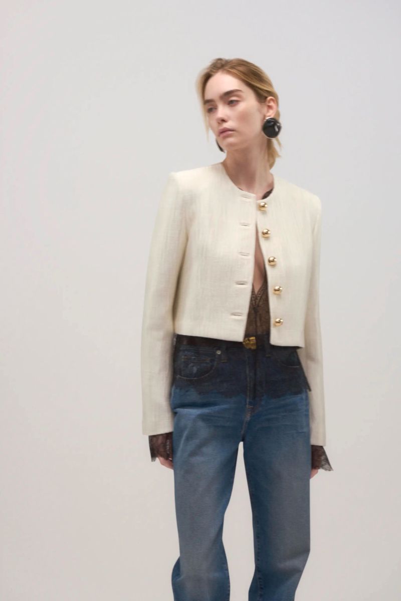 A fair-skinned adult female poses in a cream-colored jacket with gold buttons, blue jeans, and large black earrings against a white backdrop.