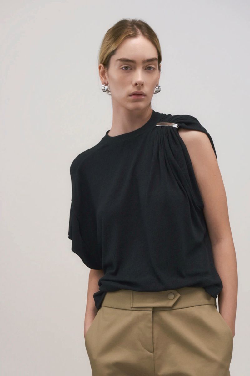 Fashion editorial image featuring a model in a black draped top and tan pants against a neutral backdrop in a studio setting.