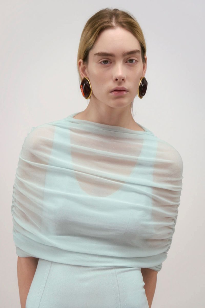 A blond woman with neutral expression wears statement earrings and a draped aqua dress against a bright neutral backdrop, studio lighting.