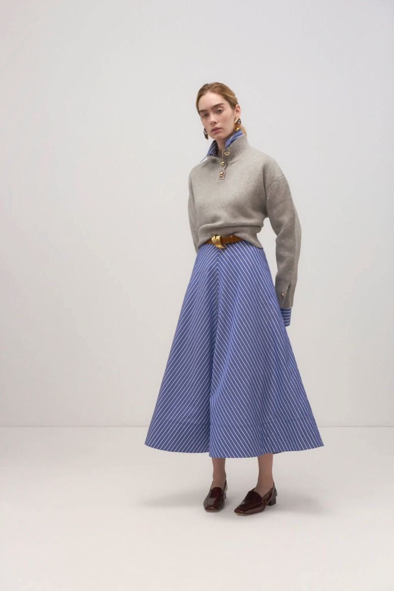 A single fair-skinned woman poses in a studio, wearing a gray sweater, blue striped skirt, brown belt, and loafers in a fashion shot.