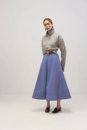 A single fair-skinned woman poses in a studio, wearing a gray sweater, blue striped skirt, brown belt, and loafers in a fashion shot.
