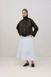 Model wearing a dark leather jacket and a white skirt standing against a plain white studio background.