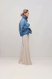 Model standing against a plain white background wearing a blue denim jacket and a long beige skirt holding a cream handbag