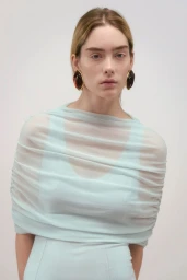 Model wearing a pale blue ruched top and dark statement earrings against a neutral grey studio background