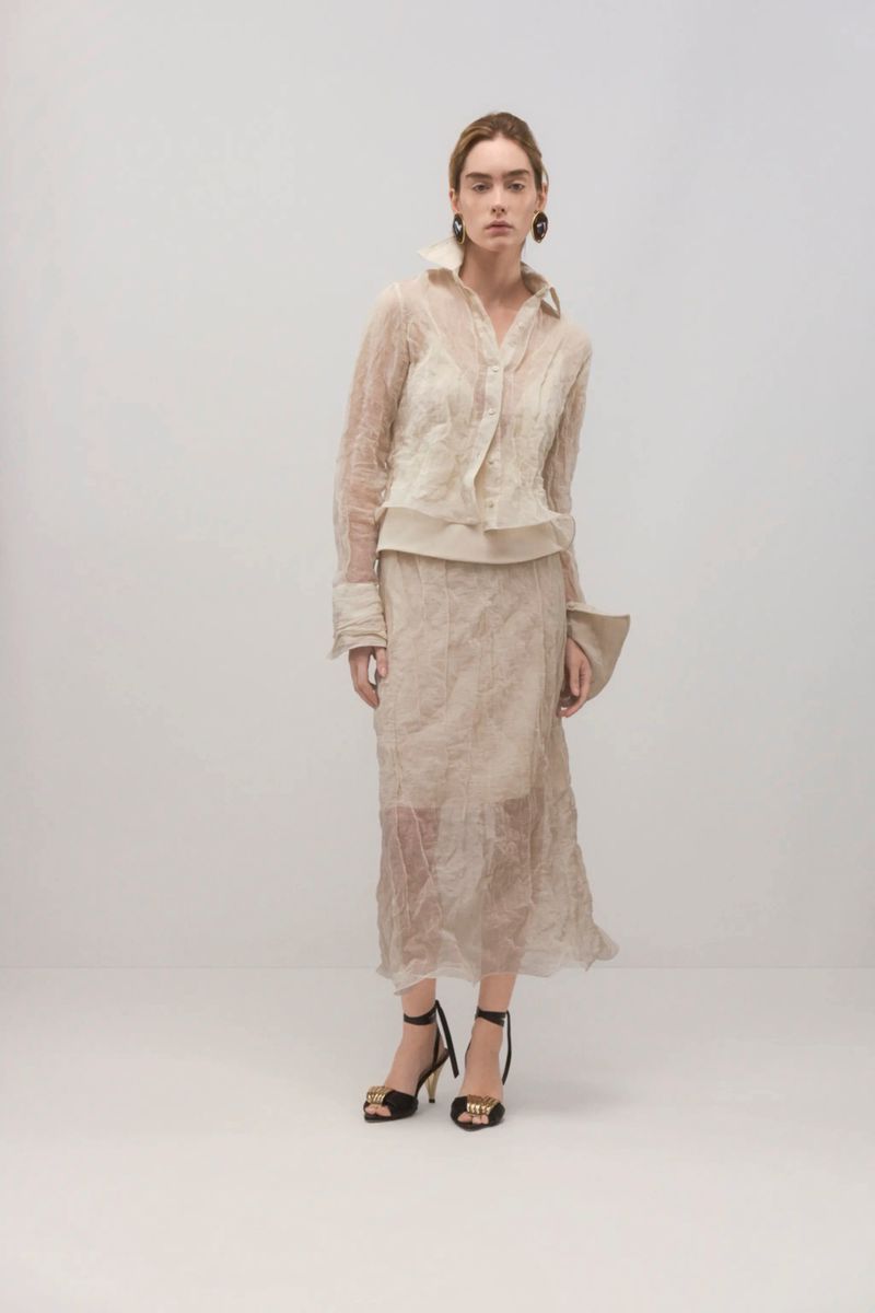 Model wearing a sheer cream-colored blouse and skirt set with black strappy heels against a neutral studio background.