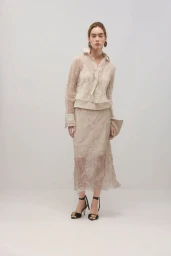 Model wearing a sheer cream-colored blouse and skirt set with black strappy heels against a neutral studio background.