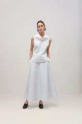 Model wearing a white sleeveless top and long white skirt standing against a neutral studio backdrop