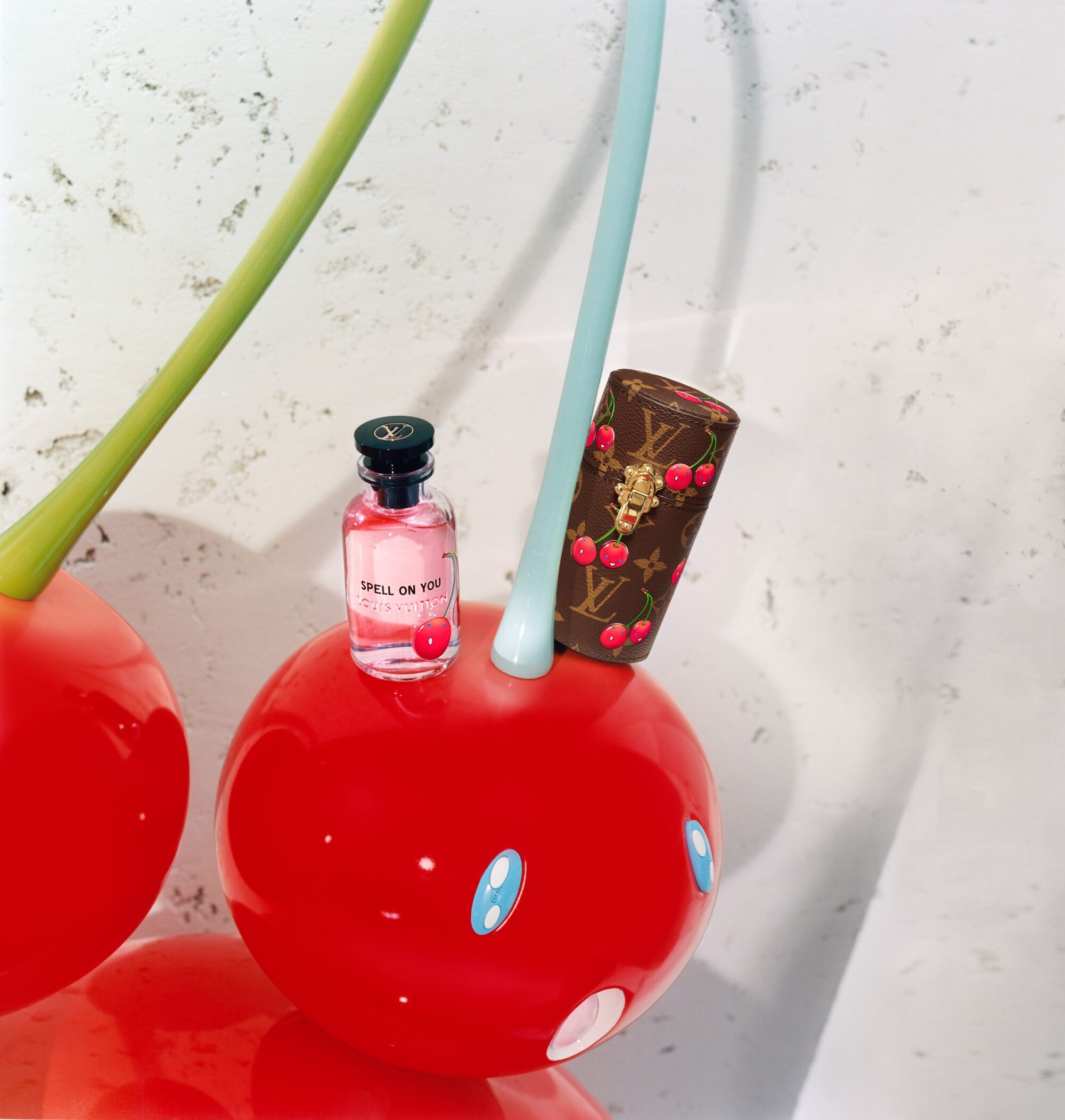 Louis Vuitton perfume bottle and monogram cylinder resting on giant red cherry sculpture with smiling blue-eyed face