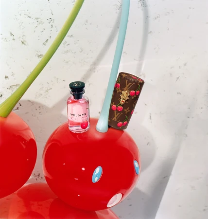 Louis Vuitton perfume bottle and monogram cylinder resting on giant red cherry sculpture with smiling blue-eyed face